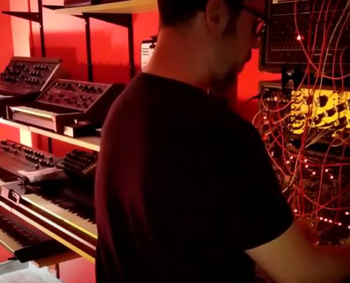 man playing synthesizer with other keyboards and synths on shelves